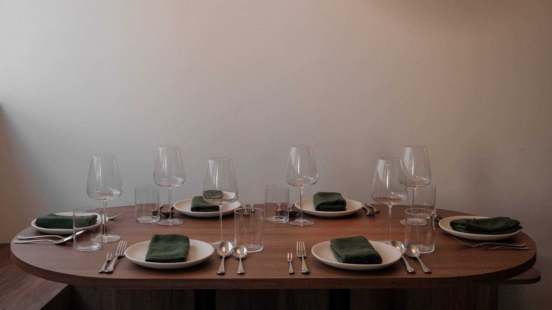 An oval wooden table set for six with white plates, green linen napkins, polished silverware, and wine glasses